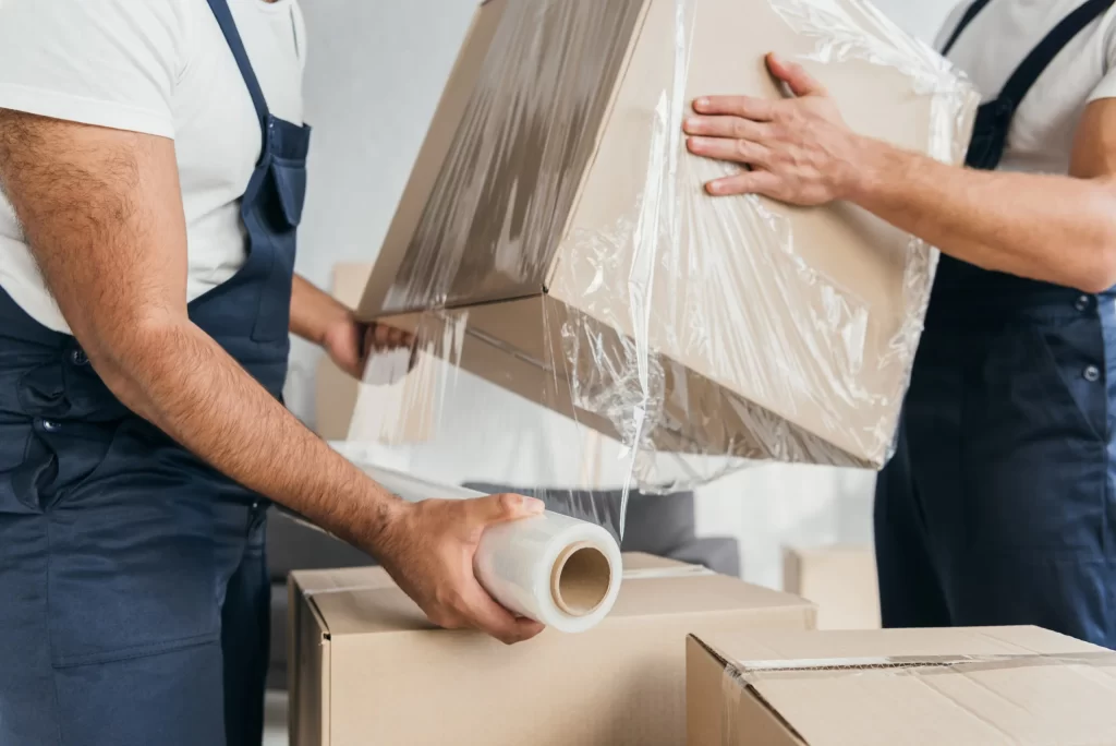 cropped view of movers in overalls wrapping box with stretch fil